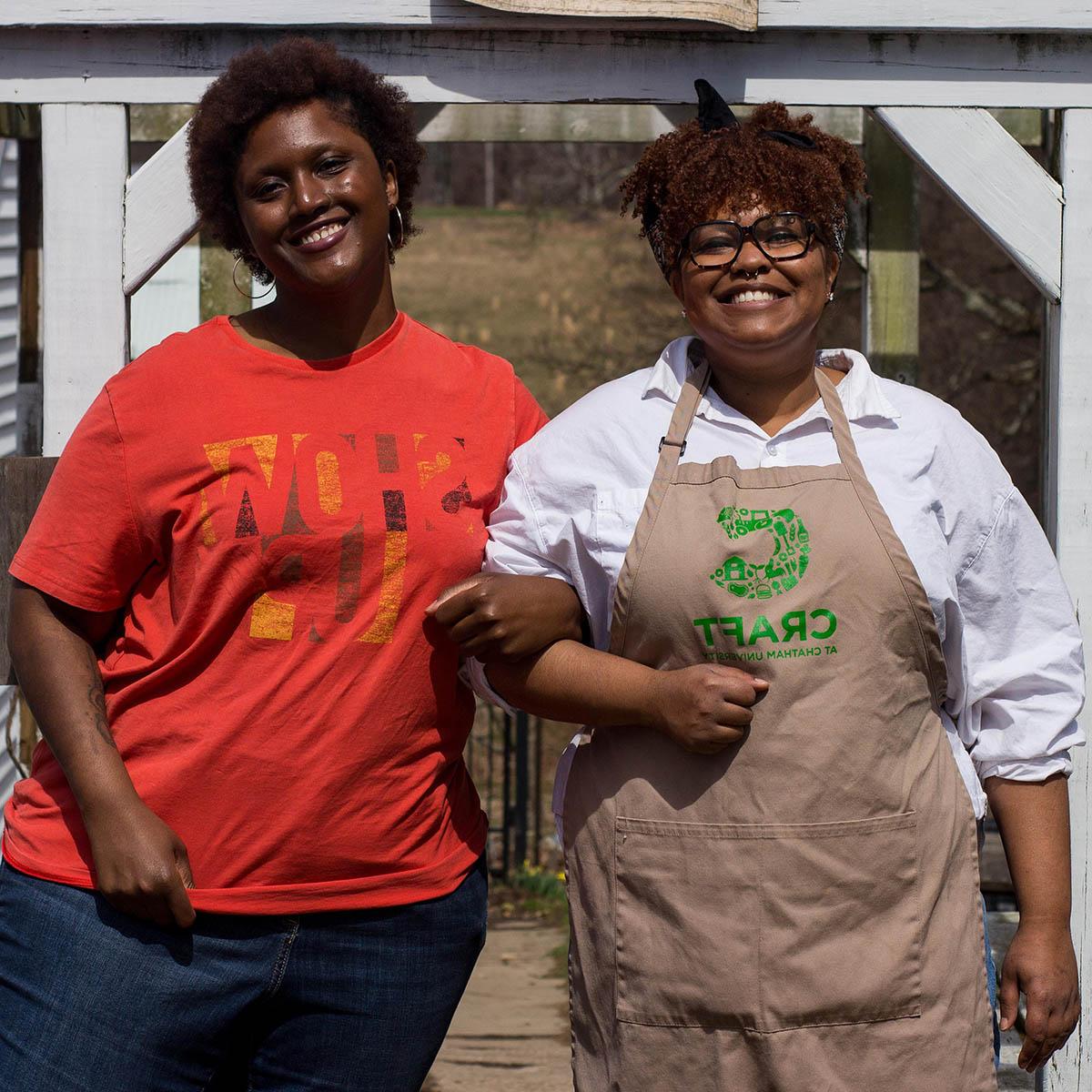 Photo of two smiling Black women linking arms. One is wearing a CRAFT apron.